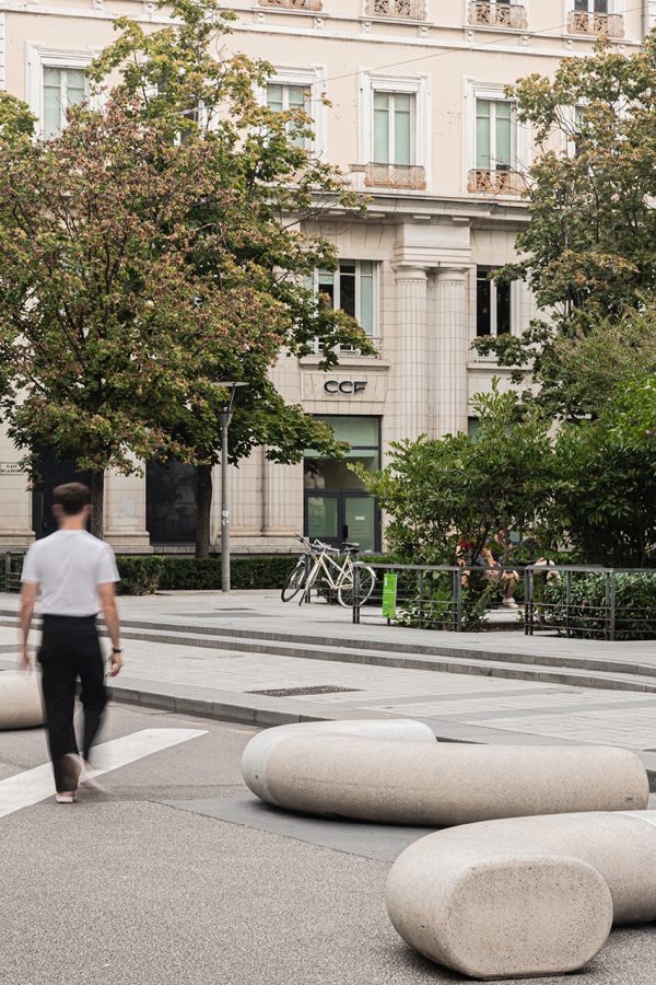 Urban installation from Durbanis’ Cinnamon collection featuring organic-shaped concrete seating modules in a tree-lined square in Lyon, with a person walking among them.